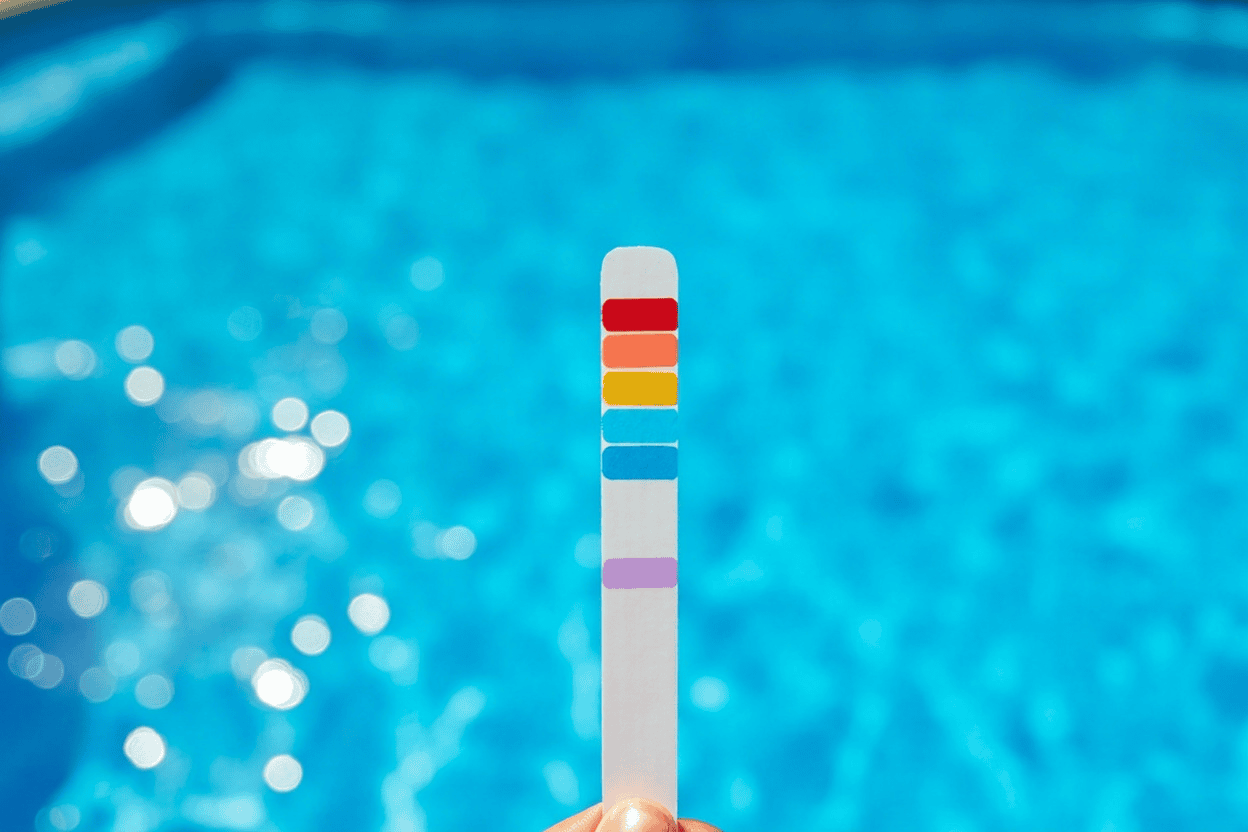 A thin paper pool test strip held up above an above-ground pool with five colored reagent patches visible